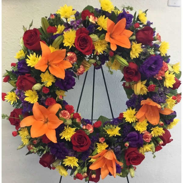 Circular wreath of red roses, orange lilies, yellow daisies, and purple flowers on a stand