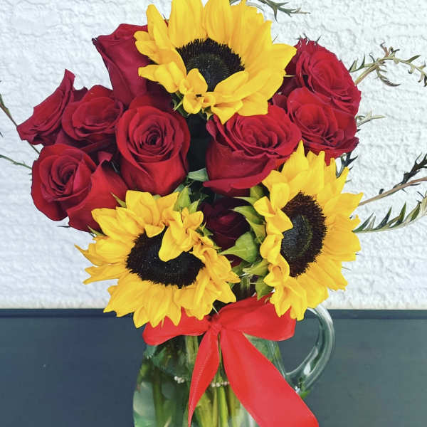 Red roses and yellow sunflowers in a clear glass pitcher with a red ribbon