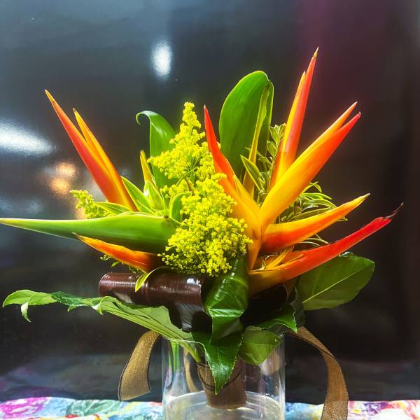 Compact tropical arrangement with orange and yellow flowers and bold green leaves in a clear glass vase.