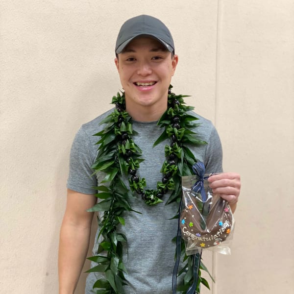 Person wearing a long green leaf lei and holding a small wrapped congratulations gift.