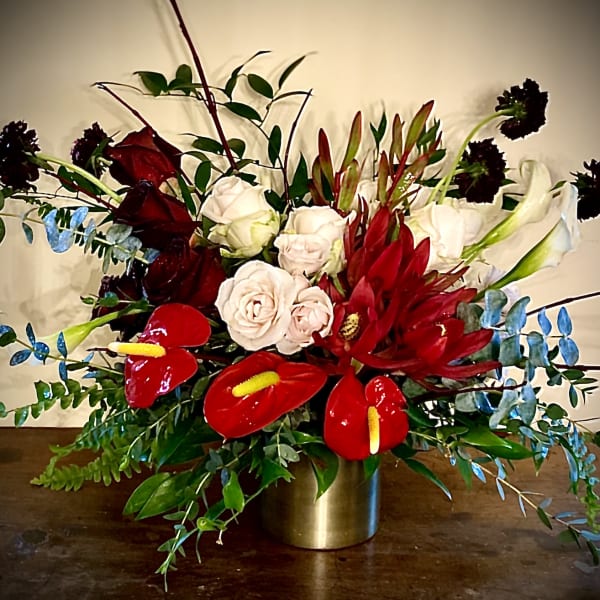 Low gold vase with red and blush roses, red tropical blooms, and white lilies in a lush arrangement.