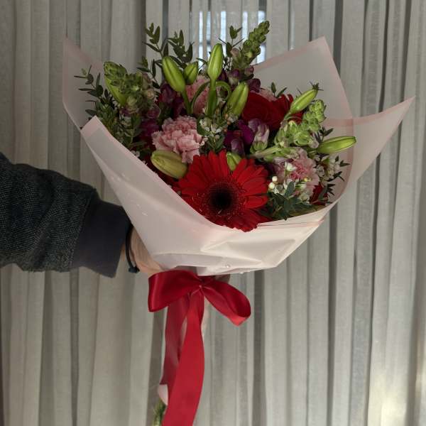 Handheld bouquet of red gerbera daisies, pink carnations, and lily buds wrapped in pale pink paper