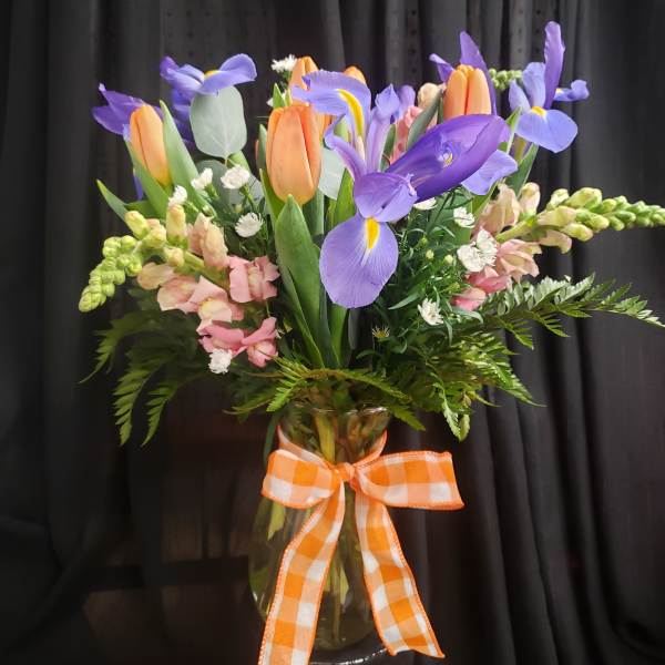 Vase of purple irises and orange tulips with pink blooms and a gingham bow.