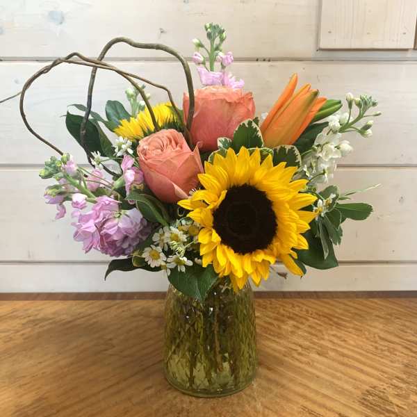 Mixed bouquet with sunflowers, peach roses, lilies, and lavender blooms in a green glass vase
