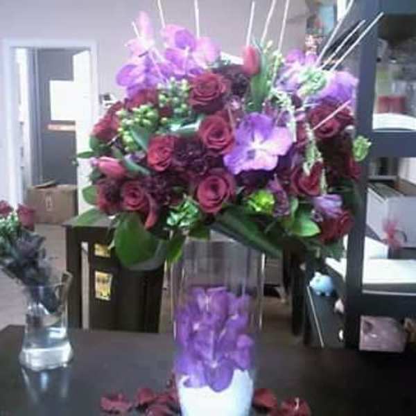 Purple and burgundy floral arrangement in a clear vase with a purple orchid cylinder centerpiece