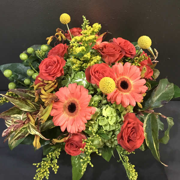 Compact arrangement of coral gerbera daisies, red roses, green hydrangea, and yellow accent flowers.
