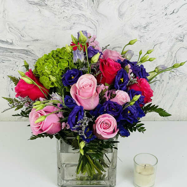 Pink and purple roses arranged in a square glass vase with a candle beside it.