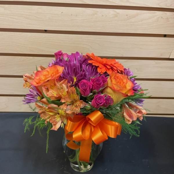 Colorful bouquet of roses, alstroemeria, and purple daisies in a glass vase with an orange ribbon