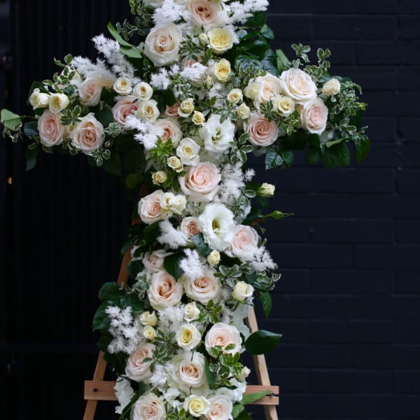 Cross-shaped floral tribute with pale roses and white blooms on an easel