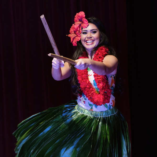 Hula dancer wearing a red flower lei and hair flower with a green grass skirt on stage