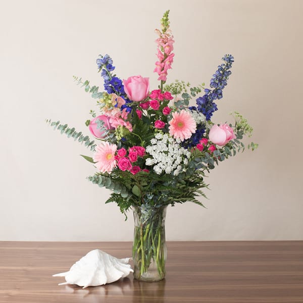 Tall pink, blue, and white mixed flower arrangement in a clear glass vase on a table