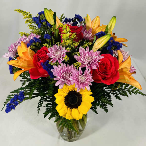 Bright mixed bouquet with sunflower, red roses, orange lilies, and purple mums in a clear glass vase
