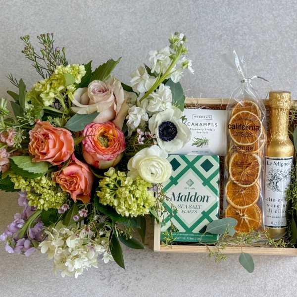 Floral bouquet beside a gift tray with caramels, sea salt, crisps, and olive oil