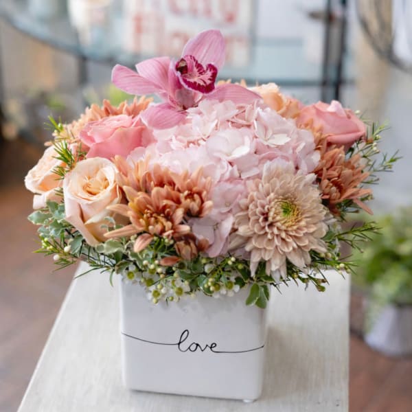 Pink and peach flowers arranged in a white square vase labeled love.