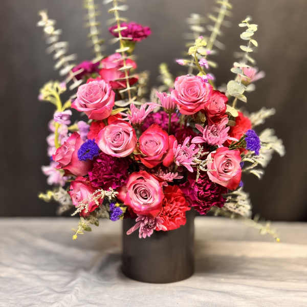Vibrant arrangement of pink roses and red carnations with purple accents in a black cylinder vase