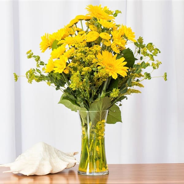 Tall bouquet of yellow gerbera daisies in a clear glass vase beside a white seashell.