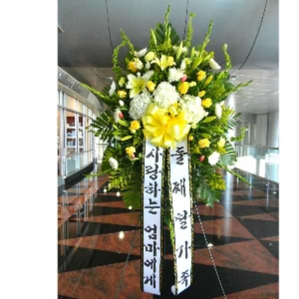 Tall standing spray of yellow and white flowers with large yellow bow and vertical message ribbons