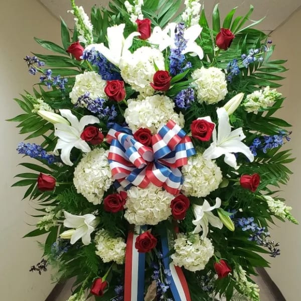 Tall standing spray of red roses, white lilies, white hydrangeas, and blue flowers with a red, white, and blue bow.
