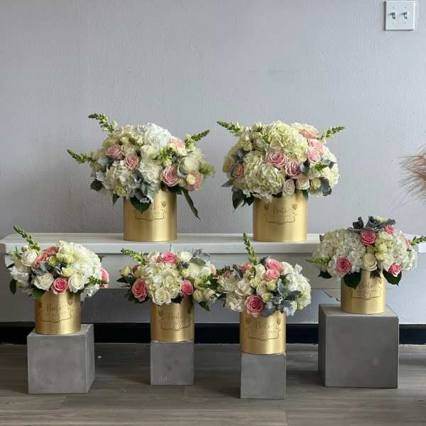Five pastel floral arrangements in gold cylindrical boxes on gray pedestals