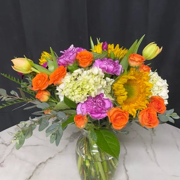 Colorful bouquet of orange roses, sunflowers, and purple carnations in a glass vase