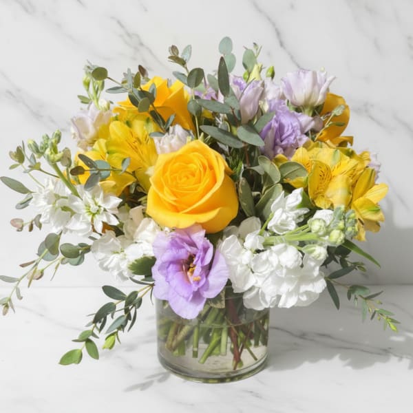 Low glass vase of yellow roses, lavender lisianthus, white stock, and yellow alstroemeria