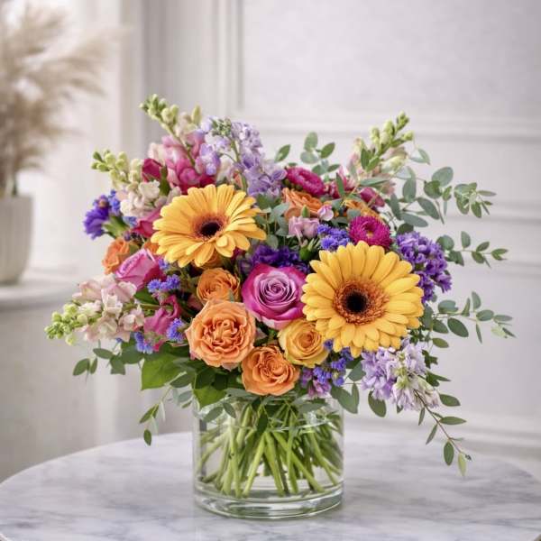Bright mixed bouquet of yellow gerbera daisies and roses in a clear glass vase on a round table.