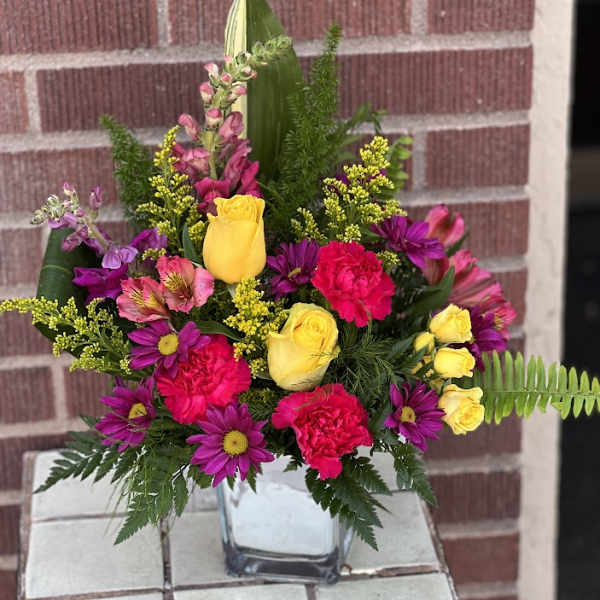 Mixed bouquet of yellow roses, pink carnations, and purple daisies in a glass vase