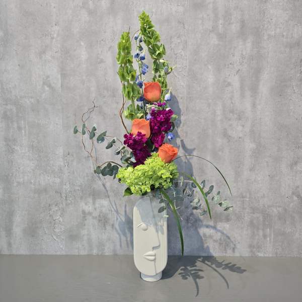 Tall floral arrangement with orange roses, purple blooms, and green hydrangea in a white vase