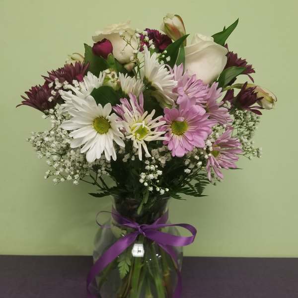 Mixed bouquet of daisies, roses, and purple blooms in a glass vase with ribbon