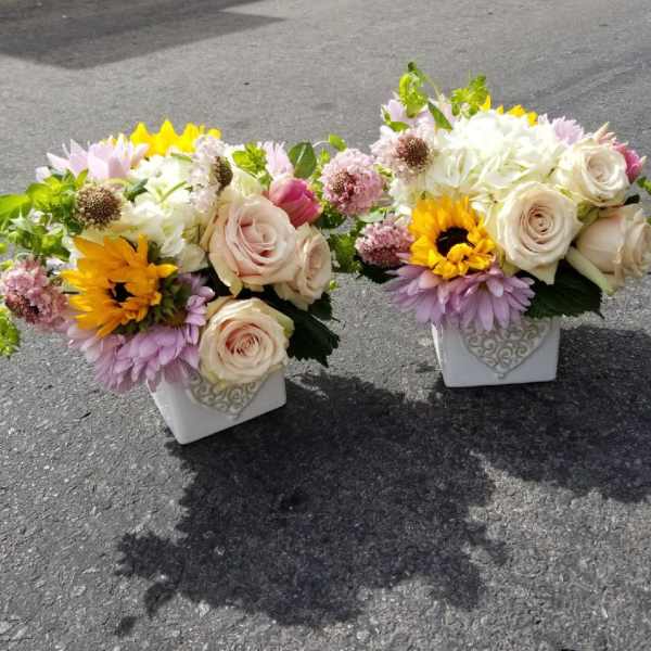 Two floral arrangements in white square vases with roses and sunflowers.