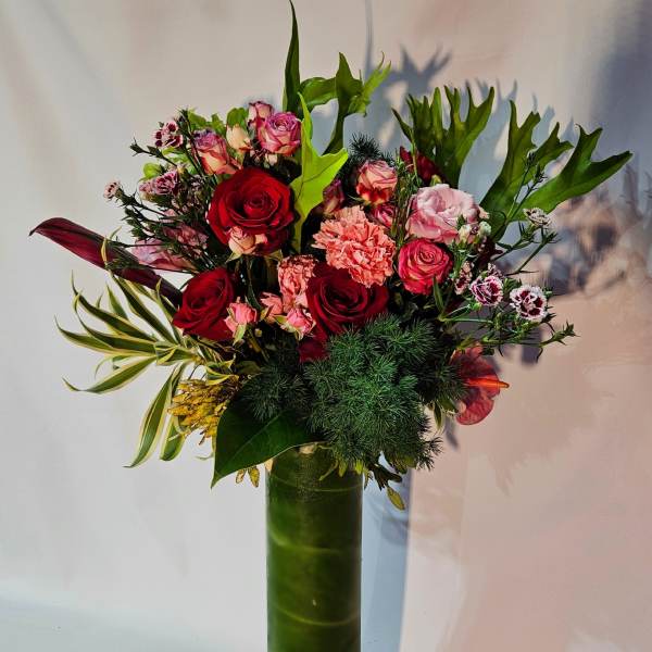 Tall arrangement of red and pink roses and carnations in a slim green glass cylinder vase