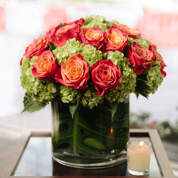 Low arrangement of orange and red roses with green hydrangeas in a glass vase beside a small lit candle.