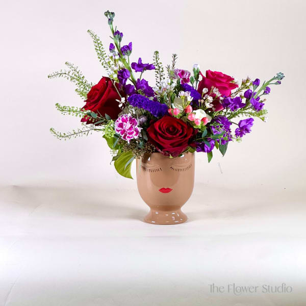 Asymmetrical arrangement of red roses and purple flowers in a tan ceramic face vase.