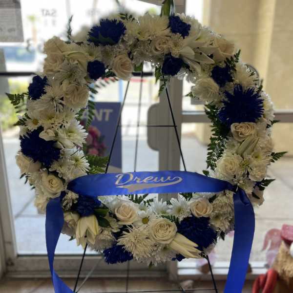 Circular floral wreath with white and blue flowers on a stand, tied with a blue ribbon