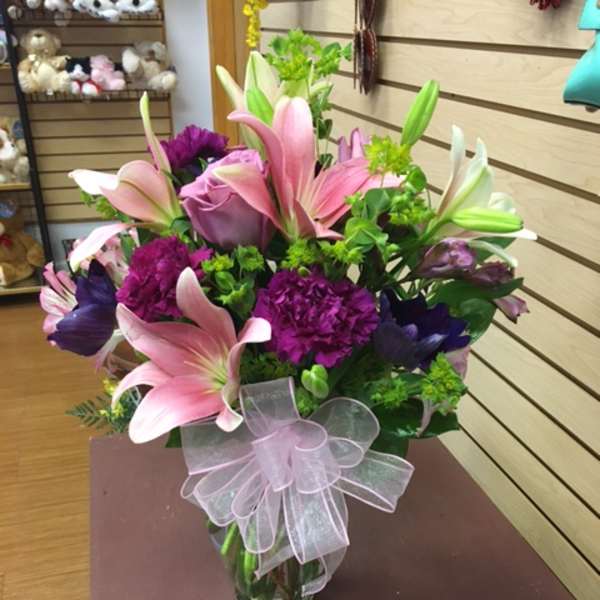 Pink lilies and purple flowers arranged in a glass vase with a sheer ribbon