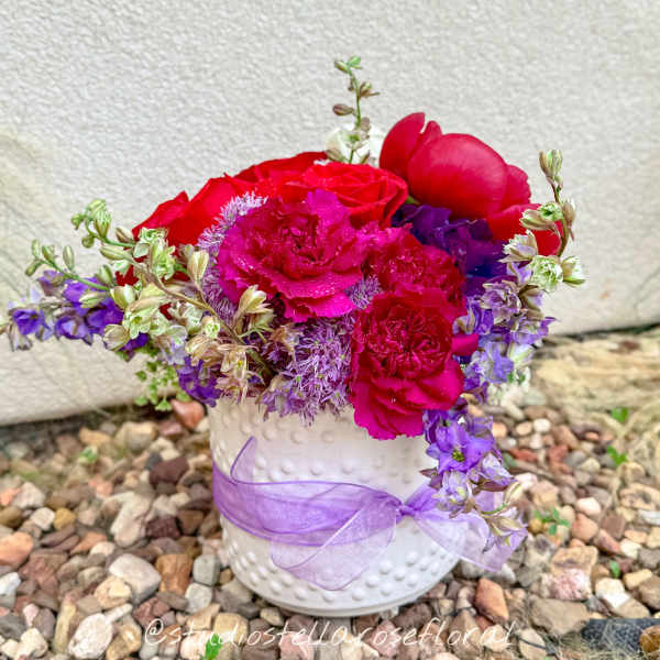 Pink and purple flowers arranged in a white vase with a lavender ribbon.