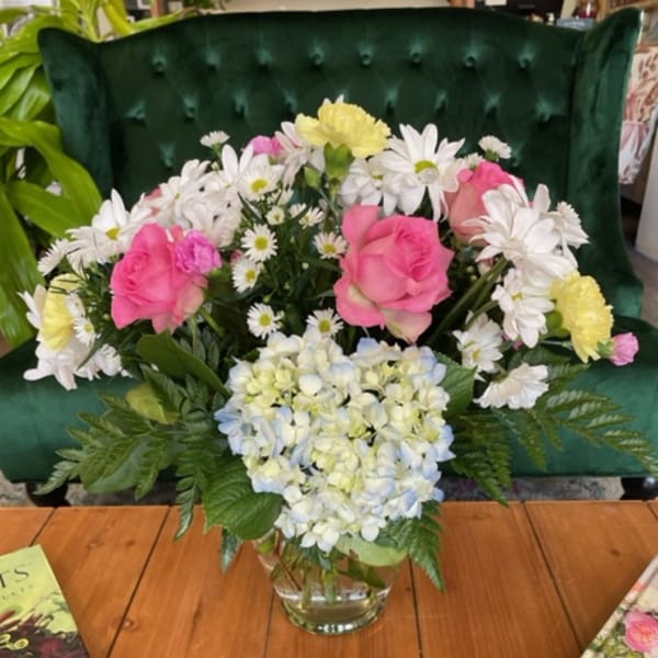 Pink roses and white daisies arranged in a glass vase with blue hydrangea