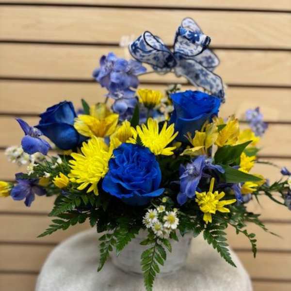 Blue roses and yellow daisies in a white vase with a patterned ribbon bow