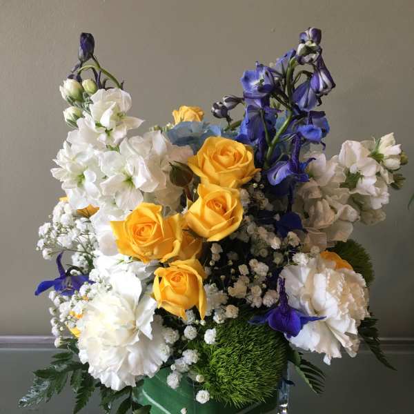 Compact arrangement of yellow roses, white blooms, and blue flowers in a square glass vase