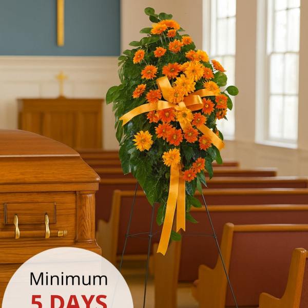 Standing spray of bright orange daisies with yellow bow on an easel beside a wood casket in a chapel.