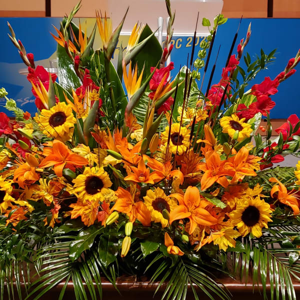 Large yellow sunflower and orange lily stage arrangement with red accents and tropical blooms.
