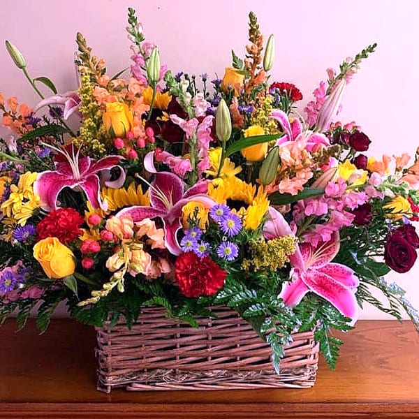 Large mixed basket arrangement with pink lilies, yellow roses, sunflowers, asters, and red carnations.