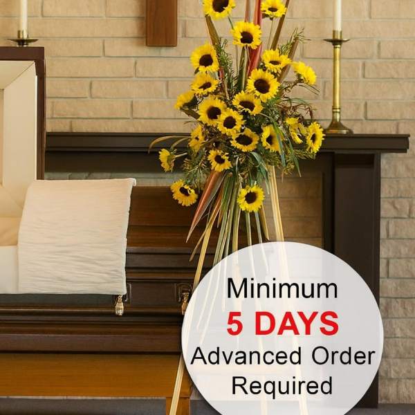 Tall easel spray of yellow sunflowers beside a wooden casket in a chapel setting