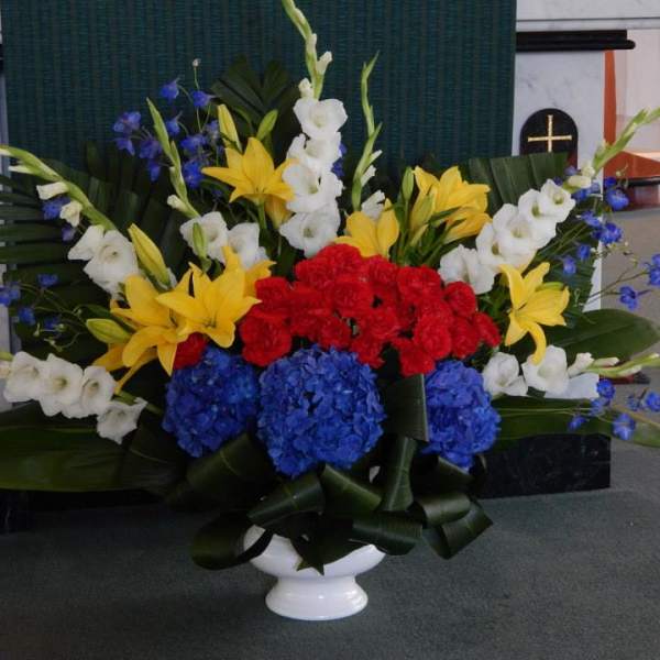 Large funeral arrangement with red, yellow, white, and blue flowers in a white vase