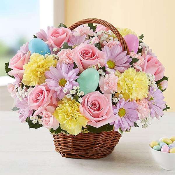 Pastel basket arrangement with pink roses, yellow carnations, lavender daisies, and decorative Easter eggs.