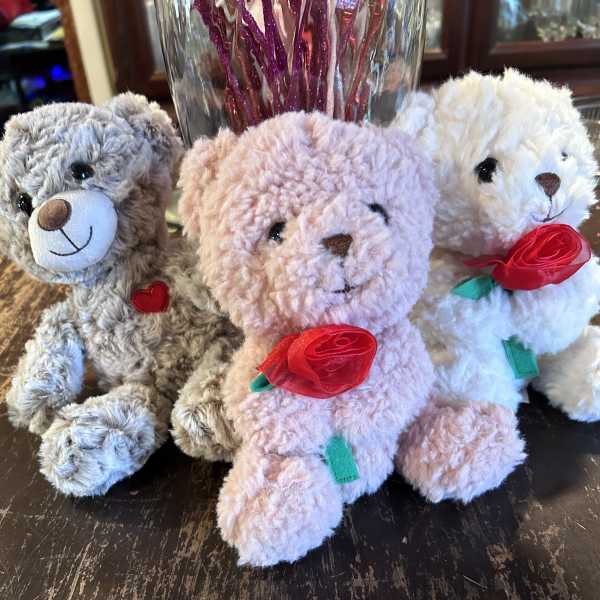 Three plush teddy bears with ribbon roses sit on a table.