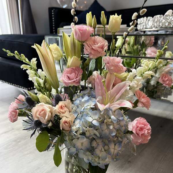 Mixed arrangement of pink lilies, roses, carnations, and blue hydrangea in a clear glass vase