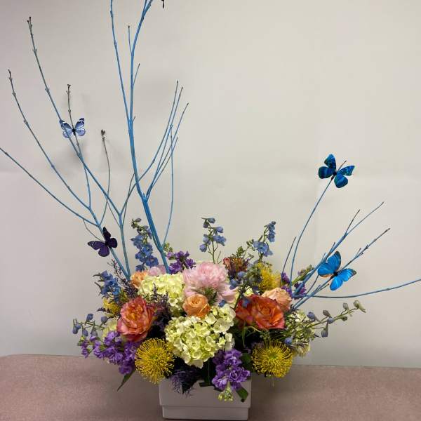 Colorful floral arrangement in a white square vase with tall blue branches and butterflies