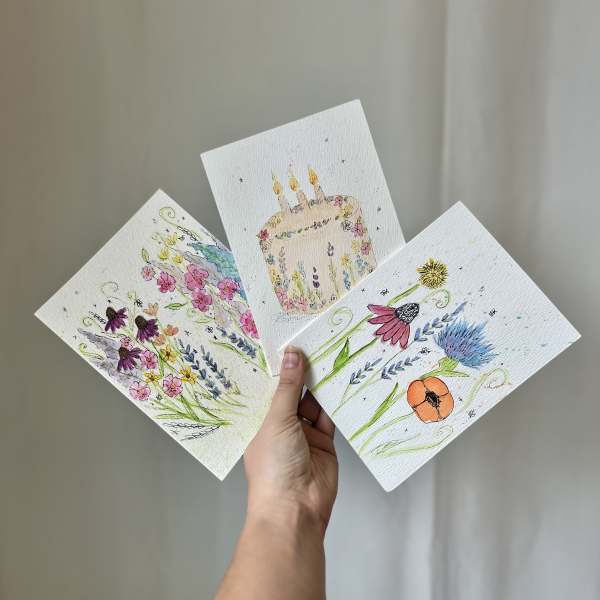 Hand holding three floral watercolor cards, including a birthday cake design