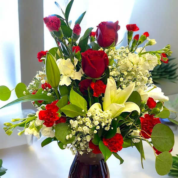 Red roses, white lilies, and red carnations arranged with filler blooms in a red glass vase.
